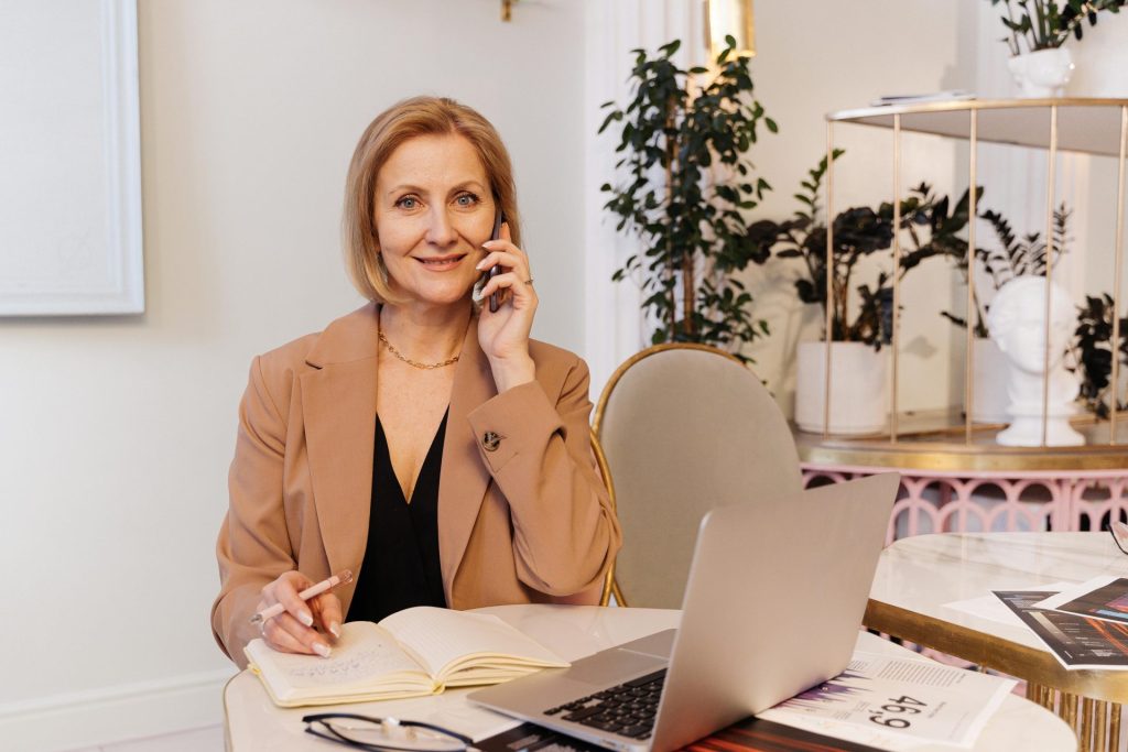 Woman in brown blazer multitasking with phone and notes at a desk.